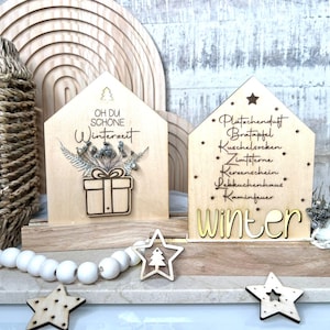 Può includere: Decorazioni in legno a forma di casa con testi e disegni a tema invernale. Uno presenta un regalo e composizioni floreali, l'altro elenca parole invernali. Sono incluse stelle di legno e una ghirlanda di perline.