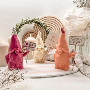 Può includere: Tre fantasiose statuette di gnomi nei toni del rosa, crema e pesca, ognuna con un cartello. Gli gnomi hanno lunghi cappelli a punta e barbe folte. Un cartello recita "Willkommen", un altro "Garten Glück", e una carta dice "Hallo Sonnenschein."