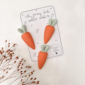 Peut inclure: Trois aimants en forme de carottes orange avec des dessus verts sont disposés sur une carte blanche. La carte comporte du texte allemand et de petites illustrations de carottes. Un objet décoratif amusant.