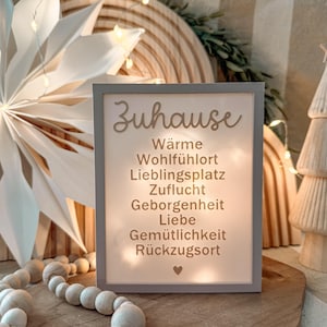 May include: A rectangular, gray-framed sign with the word "Zuhause" and other German words in a light-colored script. The sign is illuminated from behind, and the background includes a wooden star, wooden beads, and a wooden arch.