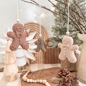 Könnte beinhalten: Drei hängende Lebkuchenmann-Ornamente in verschiedenen Brauntönen, jeweils mit einem strukturierten strickähnlichen Muster, einer Fliege und einer kleinen Holzperle an der weißen Schnur. Die Ornamente hängen an Zweigen und zeigen eine detaillierte Oberflächenstruktur, die bei jedem Körperteil gestrickten Stoff imitiert.