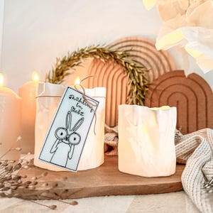 May include: Two white ceramic candle holders with a textured surface are displayed on a wooden board. One holder has a tag with a bunny illustration and the text "Frühling in Sicht". A wreath and decorative wooden arches are in the background.
