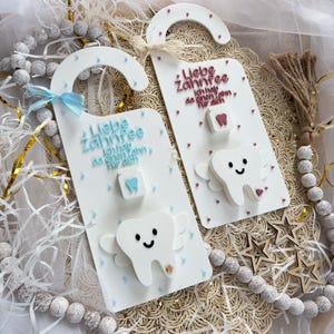 May include: Two white door hangers with a tooth fairy theme. Each hanger has a smiling tooth with wings, the text "Liebe Zahnfee" and a small tooth-shaped accent. One has a blue bow.