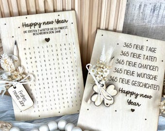 Könnte beinhalten: Zwei Holztafeln mit graviertem Text und dekorativen Elementen. Eine zeigt ein Wortsuchrätsel und den Satz „Happy New Year“. Die andere hat „365 neue Tage“ und ein Kleeblatt. Beide sind mit Trockenblumen verziert.