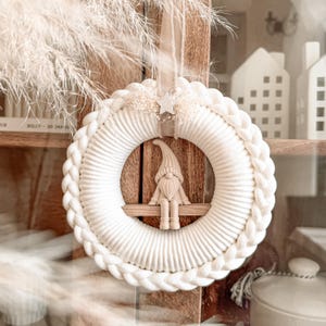 Op de afbeelding: Een ronde krans van gebroken wit gevlochten koord met een klein beige kabouterfiguurtje dat op een horizontale houten plank in het midden zit. Een kleine witte ster en gedroogde bloemen zijn aan de bovenkant van de krans bevestigd.