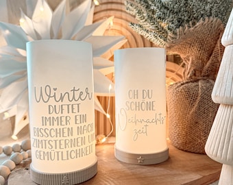 Puede incluir: Dos portavelas cilíndricos blancos con texto, "Winter duftet immer ein bisschen nach Zimtsternen und Gemütlichkeit" y "Oh du schöne Weihnachtszeit". Los portavelas están sobre una superficie de madera con una base en forma de estrella.