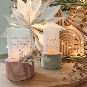 May include: Two glass candle holders with text, one on a pink base with a tree design, the other on a green base with a star. The candle holders are decorated with stars and dried flowers, with a large white paper star in the background.