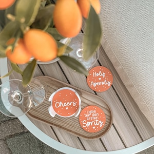 Puede incluir: Vista aérea de una bandeja de madera con tres posavasos naranjas y dos copas de vino. Los posavasos tienen texto blanco que dice "Cheers", "Holy Aperoliy" y "Liebe auf den ersten Spritz". Frutas naranjas y hojas verdes en el fondo.
