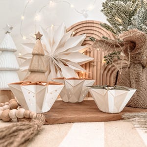 May include: Three white, geometric, star-shaped bowls with faceted sides and tan interior rims. The bowls feature small, circular cutouts along the edges and are arranged on a wooden surface alongside decorative holiday items.