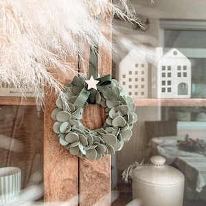 May include: A small circular wreath made of sage green eucalyptus leaves, featuring a dark green ribbon tied in a bow at the top. A small, light-colored wooden star is attached to the center of the ribbon bow, providing a decorative accent.