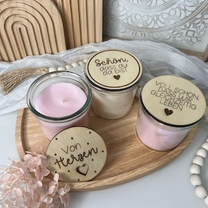 Könnte beinhalten: Ein Holztablett hält drei Glas-Kerzengläser mit Holzdeckeln und einen Holzuntersetzer. Die Kerzen sind mit rosa und cremefarbenem Wachs gefüllt. Die Deckel und der Untersetzer haben deutschen Text und Herzmotive. Dekorative Holzregenbögen sind im Hintergrund.