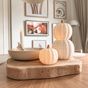 May include: A set of three white ceramic pumpkin-shaped lanterns with perforated dot patterns, featuring a stacked pair and a single smaller pumpkin, arranged alongside a shallow bowl containing a tall cream-colored candle on a thick wooden slab.