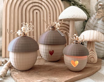 May include: Three wooden acorn-shaped decorations with brown caps and light-colored bodies. Each has a small red heart detail. The acorns are adorned with dried floral accents and sit on a wooden tray, creating a rustic, nature-inspired theme.