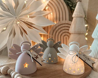 Puede incluir: Tres pequeñas figuras de ángeles en colores pastel suaves, cada una con un recorte en forma de estrella que brilla desde el interior. El ángel blanco de la derecha tiene una etiqueta de madera con el texto "Glücks LICHT" y una estrella grabada.