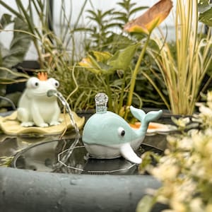 Puede incluir: Una fuente de agua solar con forma de ballena en color verde claro y blanco con un pequeño chorro de agua que brota de la parte superior, colocada en un cuenco de agua oscuro junto a una figura de rana blanca con una corona naranja.