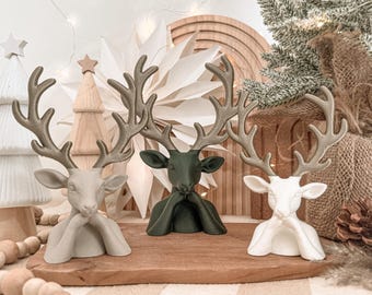Puede incluir: Tres figuras de ciervo con grandes astas, cada una de un color diferente: gris claro, verde oscuro y blanco. Cada ciervo está representado en estilo busto con sus pezuñas delanteras colocadas cerca de la barbilla, mostrando un acabado liso.