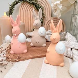 Puede incluir: Tres figuras de conejos de Pascua en colores pastel, cada una sosteniendo un huevo blanco con un diseño de corazón. Los conejos son en tonos rosa, beige y melocotón. El fondo incluye velas, arcos de madera y elementos decorativos.