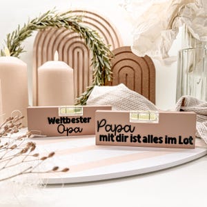 Op de afbeelding: Twee rechthoekige, lichtbruine waterpassen met zwarte tekst. Op één staat "Weltbester Opa" en op de andere "Papa mit dir ist alles im Lot". Elke waterpas heeft een kleine, transparante waterpas. De items liggen op een gestreept oppervlak.