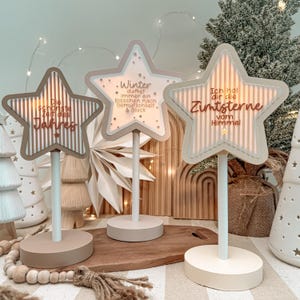 Puede incluir: Tres decoraciones de mesa en forma de estrella sobre postes blancos y bases redondas. Cada estrella presenta rayas verticales y texto en alemán: Die schönste Zeit des Jahres, Winter duftet immer ein bisschen nach Gemütlichkeit & Glück, e Ich hol dir die Zimtsterne vom Himmel.
