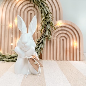 May include: A white ceramic bunny figurine with a tag tied around its neck. The bunny has long ears with dotted details and is holding its paws together. A wooden arch and a green wreath are in the background.