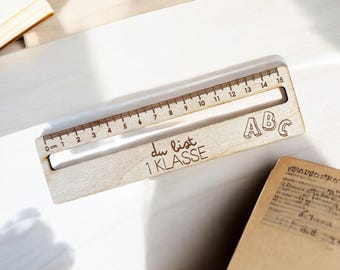 Puede incluir: Una regla de madera con una escala de 15 cm, el texto "du bist 1 KLASSE" y las letras "ABC" grabadas en ella.