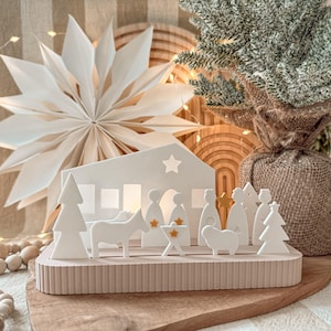 May include: A white Christmas nativity scene with a house, figures, and animals. The scene is set on a light pink base. A large white paper star and a small Christmas tree are in the background. The scene is on a wooden board.