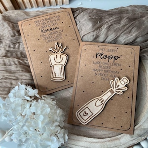 Puede incluir: Dos tarjetas marrones con un tapón de botella de madera y un corcho de madera que sale de una botella. El texto en las tarjetas dice "Dein Geburtstag ist der perfekte Grund, den Korken mal wieder zu zeigen wo's langgeht!" y "Mit jedem Plopp wird das Leben besser - Happy Birthday & Prost auf Dich!"