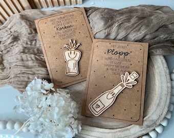 May include: Two brown cards with a wooden bottle stopper and a wooden cork popping out of a bottle. The text on the cards reads "Dein Geburtstag ist der perfekte Grund, den Korken mal wieder zu zeigen wo's langgeht!" and "Mit jedem Plopp wird das Leben besser - Happy Birthday & Prost auf Dich!"