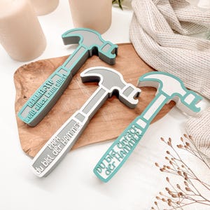 Peut inclure: Trois ouvre-bouteilles en forme de marteau, gris, blanc et turquoise. Chaque ouvre-bouteille porte une phrase allemande différente imprimée sur le manche. Les ouvre-bouteilles sont disposés sur une planche à découper en bois, avec un fond neutre.