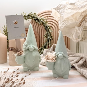 Könnte beinhalten: Zwei mintgrüne Zwergfiguren mit konischen Hüten und langen Bärten. Jeder Zwerg hält einen kleinen Topf mit Trockenblumen. Eine Karte mit einer Biene und den Worten "Bee Happy" ist in der Nähe aufgestellt. Der Hintergrund umfasst Kerzen und dekorative Elemente.