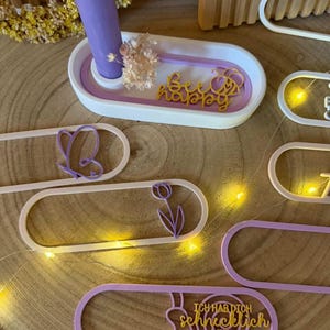 Puede incluir: Un portavelas decorativo con una vela morada, flores secas y un letrero dorado "Bee happy". También hay varias decoraciones de forma ovalada con diseños recortados, incluyendo una mariposa, un tulipán y texto en alemán. Los artículos son en tonos blanco, morado y dorado.