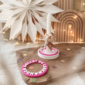 Könnte beinhalten: Eine weiße Kerze mit floralem Akzent steht auf einer rosa-weißen dekorativen Basis. Ein rosa Ring mit den Worten "SOLLST DU LEBEN HOCH" liegt daneben. Die Artikel befinden sich auf einem Holzbrett mit Konfetti und einem Papierstern im Hintergrund.