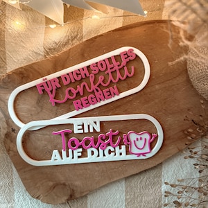 Puede incluir: Dos letreros blancos de forma ovalada con texto rosa sobre una superficie de madera. El letrero superior dice "FÜR DICH SOLL ES Konfetti REGNEN". El letrero inferior dice "EIN Toast AUF DICH" con un gráfico de tostada sonriente.