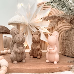 May include: Three squirrel figurines in varying shades of brown and beige, each featuring a textured knit-like surface pattern. Two of the squirrels are holding small acorns in their front paws, while the third stands upright with paws tucked.