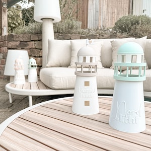 May include: A collection of pastel-coloured lighthouse ornaments, including white, pink, and mint green, are arranged on a wooden table. The lighthouses feature detailed designs, with the words "Heimat Hafen" and "Nordlicht" inscribed on two of them.