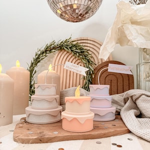 Può includere: Una collezione di candele decorative a forma di torta in colori pastello, con un design a glassa colante. Una candela ha una fiamma gialla e alcune hanno delle bandierine con le scritte "Just Married" e "Pustekuchen". Le candele sono su una tavola di legno.