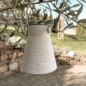 Könnte beinhalten: Eine weiße, kegelförmige Außenlaterne mit Metallgriff, die an einem Ast hängt. Die Laterne trägt geprägten Text auf Deutsch, darunter das Wort "Sommer" und andere sommerbezogene Wörter. Der Hintergrund zeigt eine verschwommene Außenszene.