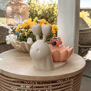May include: A whimsical, off-white ceramic snail figurine with heart-shaped antennae and a peach-colored flower-shaped dish. A small ladybug figurine rests in the dish. The snail is on a round, light-colored wooden table.