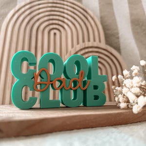 Pode incluir: Uma placa turquesa "COOL CLUB" com uma sobreposição laranja "Dad", colocada sobre uma superfície de madeira. O fundo apresenta um design de arco de madeira e um pequeno buquê de flores brancas. A placa é um item decorativo.