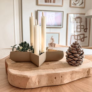 May include: A star-shaped beige candle holder containing four cream-colored taper candles and green foliage, positioned next to a brown pinecone on a thick, light-colored wooden slab with a natural, uneven edge and visible wood grain texture.