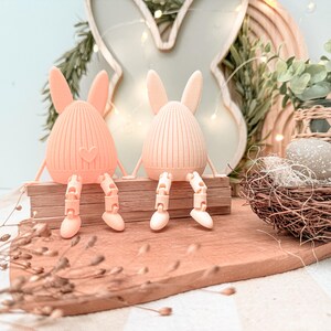 May include: Two peach-colored bunny figurines with articulated legs sit on a wooden surface. One bunny has a heart detail. The background includes a bunny-shaped cutout, greenery, and a decorative nest, suggesting an Easter theme.