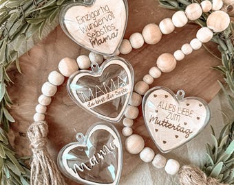 May include: Three clear heart-shaped ornaments with wooden inserts. The ornaments have different messages engraved on them: "Einzigartig Wundervoll Selbstlos Mama", "MAMA du bist die beste", and "ALLES LIEBE ZUM Muttertag".