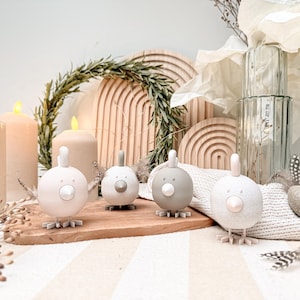 Könnte beinhalten: Vier dekorative Vogelfiguren in Weiß- und Grautönen sind auf einem Holzbrett angeordnet. Die Vögel haben ein minimalistisches Design mit rundem Körper, kleinem Schnabel und Kamm. Der Hintergrund umfasst Kerzen, Holzbögen und dekorative Elemente.