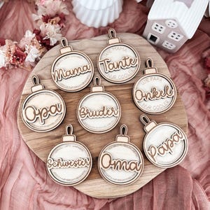 Può includere: Ornamenti natalizi in legno con nomi come "Mama", "Tante", "Opa", "Bruder", "Schwester", "Oma", "Papa" e "Onkel" su una tavola di legno. Gli ornamenti hanno una finitura in legno chiaro e sono disposti su un tessuto rosa.