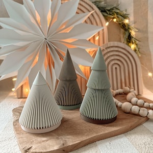 Op de afbeelding: Drie decoratieve papieren kerstbomen in groen- en bruintinten, met een witte sterrenuitbarsting decoratie op de achtergrond. De bomen zijn gerangschikt op een houten dienblad met een snoer van houten kralen.