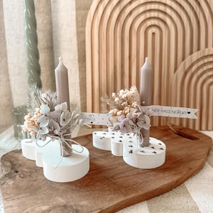Pode incluir: Dois castiçais brancos com um design em forma de coração e bolinhas pretas. Cada castiçal tem uma vela cinza e um buquê de flores secas. Um castiçal tem uma placa que diz "Hey Hase" e o outro diz "Liebster Hasengruß".