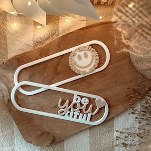 以下が含まれることがあります： 切り絵のデザインが施された、白の装飾的なブックマーク風のオブジェクトが2つ。1つは「GLÜCKLICH」と「RUNDUM」の文字が入った笑顔の顔が特徴です。もう1つはハート付きの「be you tiful」と書かれています。どちらも木製の表面にあります。