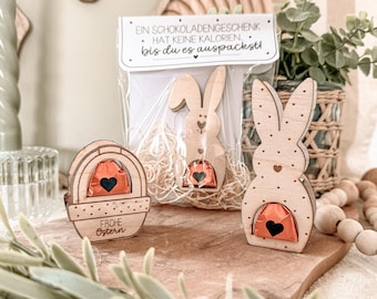 Könnte beinhalten: Drei hölzerne Osterhasen- und Korbhalter mit jeweils einem Schokoladenei. Die Hasenhalter haben ein Punkte-Muster und einen Herz-Ausschnitt. Der Korbhalter sagt "Frohe Ostern".