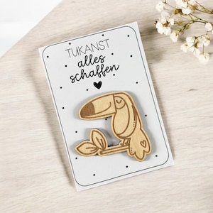 May include: A white card with black text that reads "TUKANST alles schaffen" with a small black heart. A wooden toucan with a small heart on its chest is attached to the card.