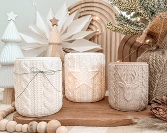 May include: Three cylindrical candle holders with a textured knit pattern. The left holder is white with a thin green string tied in a bow. The centre holder is off-white with a raised star design. The right holder is beige with a raised deer head.